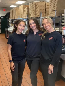 Three women in black shirts, Genova's To Go managers, standing in a pizza kitchen, preparing delicious pizzas.